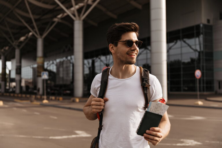 Portrait of handsome man in white tee and sunglasses holds backpack, passport and tickets and poses